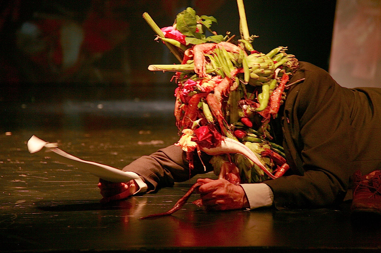Man in suit, covered in food, lies on stage; ArtBombUK