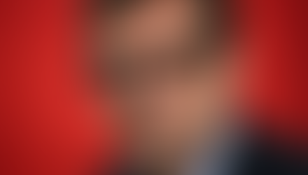 Close-up portret van een man met een serieuze blik die recht in de camera kijkt, draagt een zwarte Ray-Ban Meta smartbril tegen een opvallende rode achtergrond.