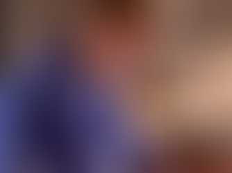Close-up of a home healthcare nurse in blue scrubs sitting at a table, focused on writing notes beside a laptop and a mug, suggesting administrative work or documentation after a shift.