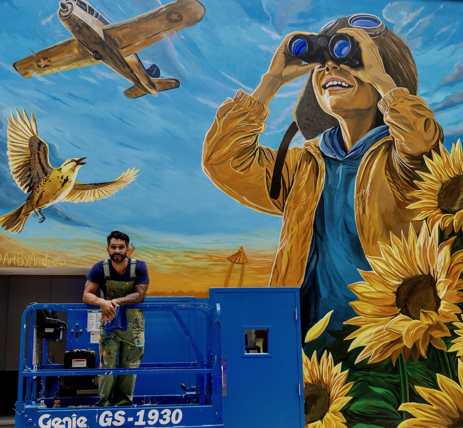Artist standing on a lift in front of the completed Wichita aviation and sunflower mural