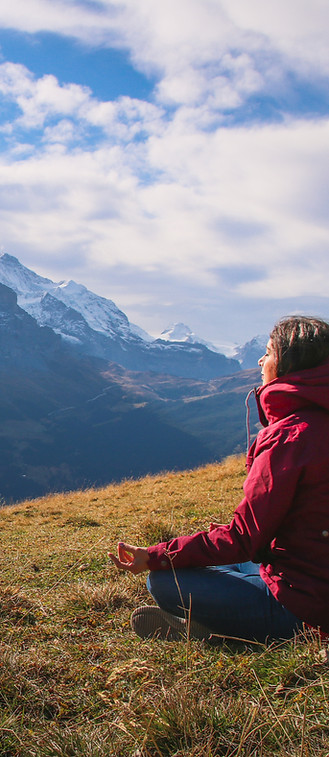 A woman in a red jacket meditating on a grassy hill facing snowy mountains, evoking deep clarity and mental peace.