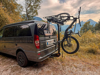 Es ist ein silberner Mercedes VIANO Marco Polo zu sehen. Am Heck ist ein schwarzer Vertikaler Fahrradträger der Firma NEOMOTUS zu sehen. Ein Fahrrad ist schon montiert. Es sieht super stimmig und stabil aus. Im Hintergrund ist der Berg Watzmann zu sehen. Das Fahrzeug steht am Waldrand und das Gras ist hellbraun bis  dunkel grün, Herbststimmung.
