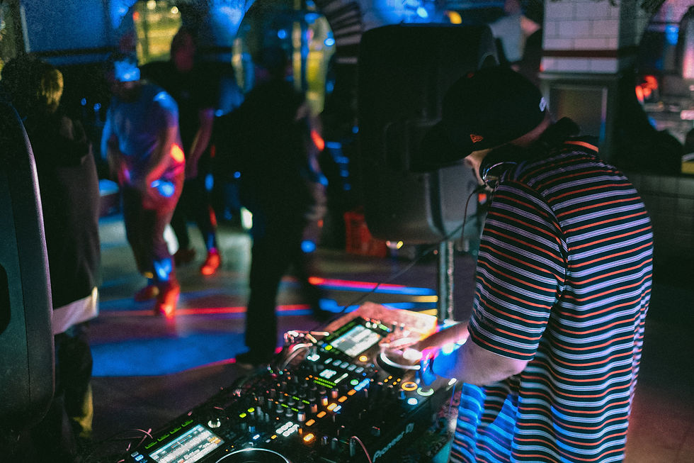 DJ Assault in striped shirt mixes music on turntables in a club. People dance in the dim, colorful light. Energetic party atmosphere. 36th Chamber