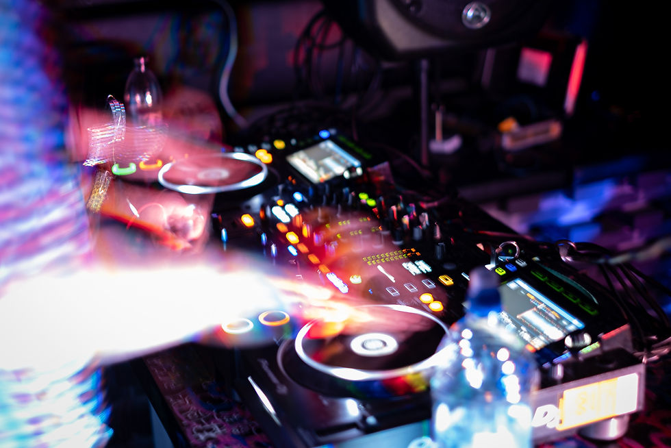 DJ Assault mixing music on a colorful, illuminated console in a dimly lit club. Motion blur highlights dynamic movement and energy.