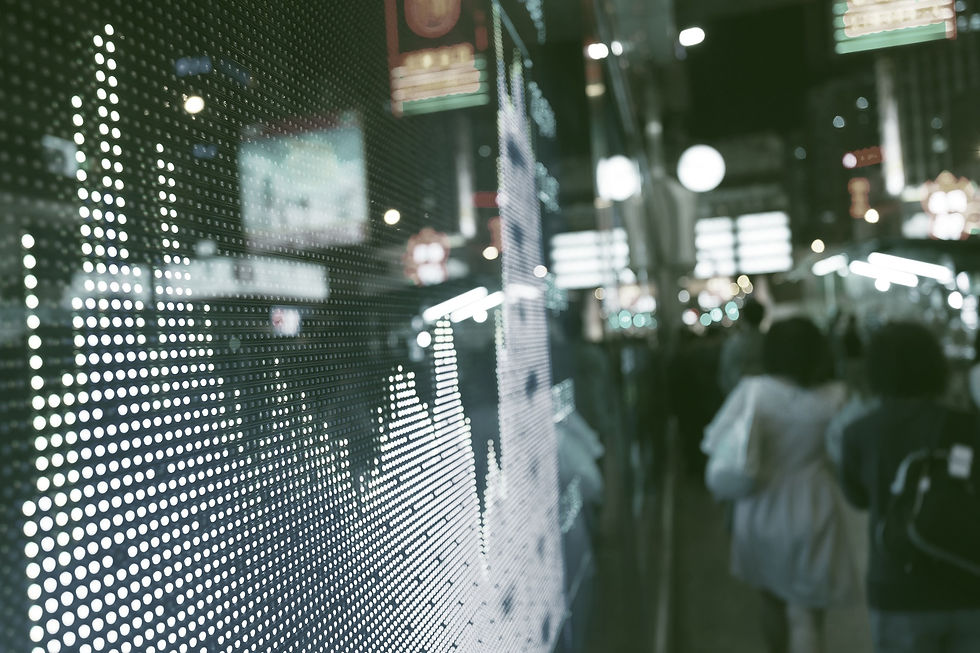 An illuminated board shows stock market movements to passerby on a crowded city street. Lights from the buildings and passing cars are reflected against the windows. Image Credit: Wix
