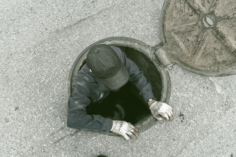 A worker wearing a uniform and industrial gloves climbs out of a sewer through an open manhole.