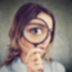 Woman holding magnifying glass over eye, enlarging it. Background is gray and textured. She wears a white shirt, showing curiosity.