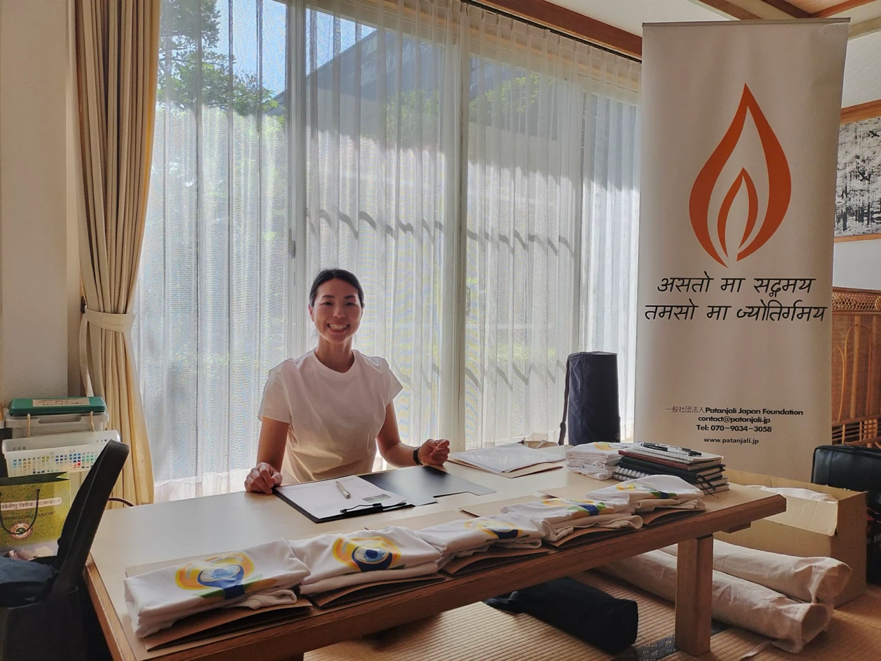 Tシャツとチラシが並ぶ受付テーブルで笑顔のスタッフが対応しています。Reception setup with a smiling staff member seated at a table, displaying t-shirts and flyers.