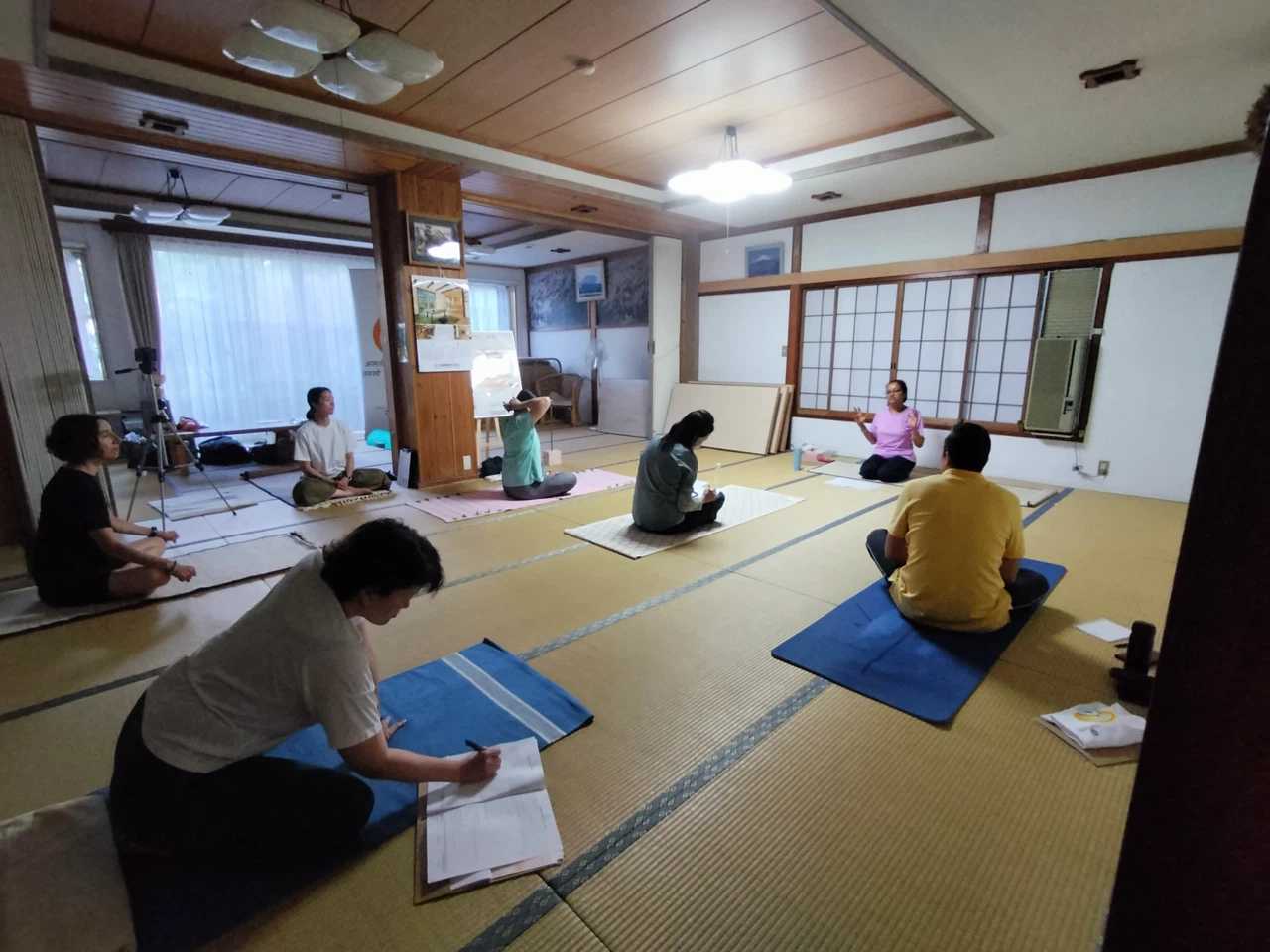 ヨガマットに座りながらメモを取る参加者たちの室内ヨガワークショップ。Indoor yoga session with participants seated on yoga mats, taking notes during a workshop.