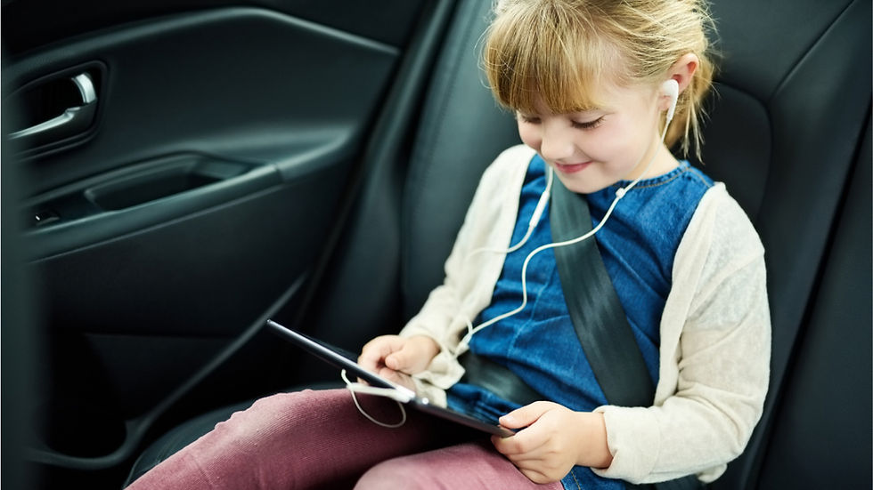 A smiling girl in a car seat watches a tablet with white earphones. She's wearing a blue dress, pink pants, and a beige cardigan.