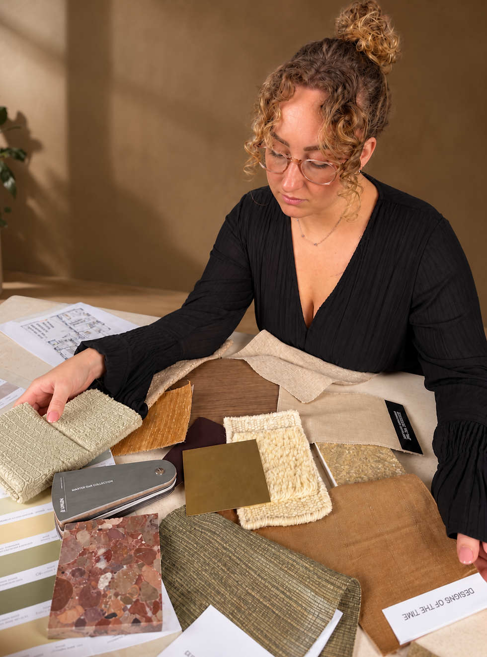 Studio photography of a woman carefully examining a variety of fabric and material swatches spread across a table. She is selecting options for a design project.