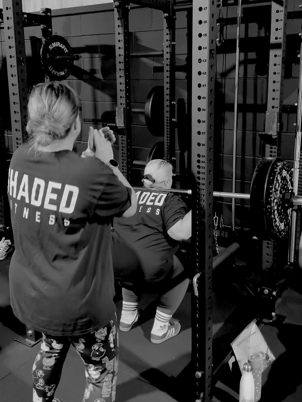 Gym member cheering on another member performing a barbell back squat at Shaded Fitness