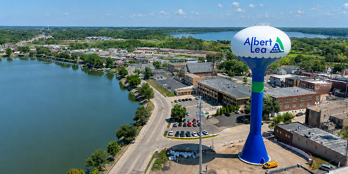 Aerial view of Albert Lea, Minnesota featuring the Albert Lea water tower, lakefront, downtown buildings, roads, trees, and surrounding lakes on a clear sunny day.