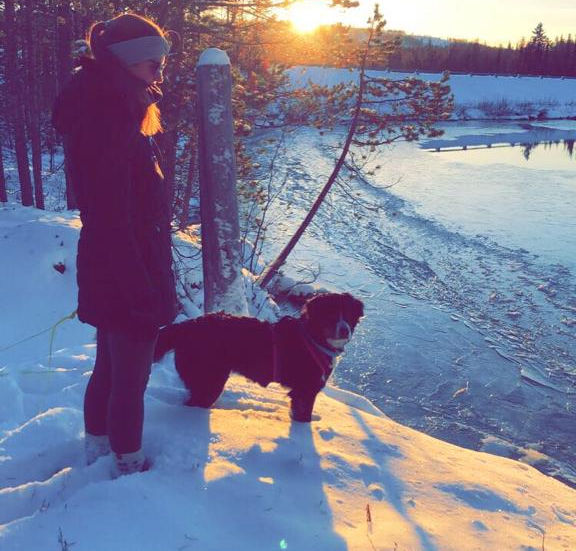 Eine Frau steht im Sonnenuntergang vor einem gefrorenen See. Ein Berner Sennenhund steht direkt neben ihr.