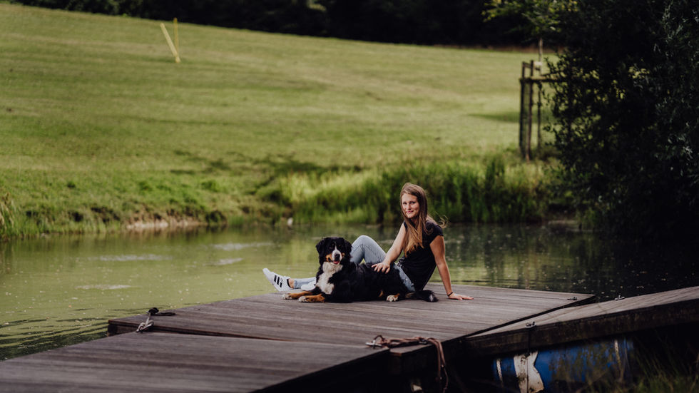 Wilma, eine junge Frau, sitzt mit ihrem Berner Sennenhund auf einem Holzsteg an einem Teich. Der Hund liegt.