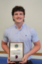 Smiling person holding a "Student of the Month" plaque. Background is plain, wearing a blue patterned shirt. Plaque text visible.