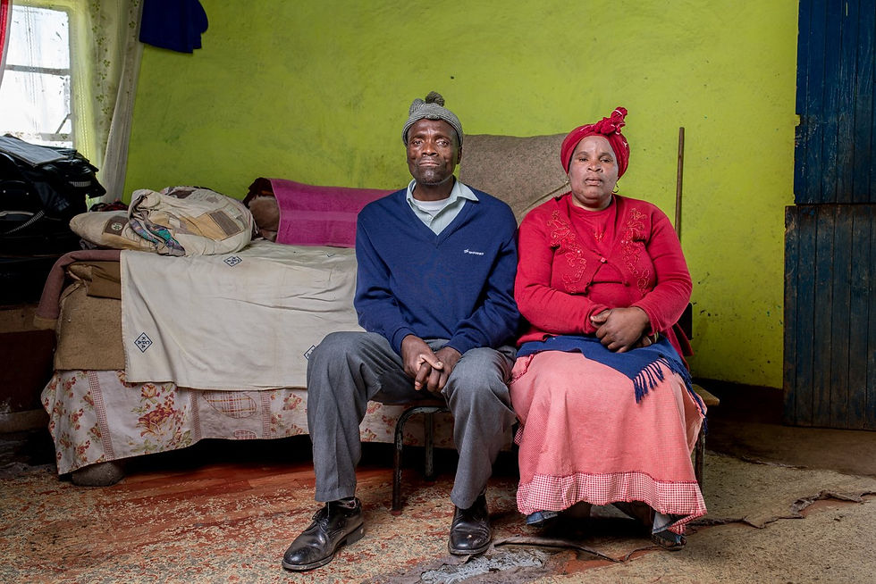 A man and woman sit together on a stool in their bedroom.