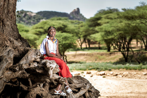 A young woman in Kenya sits on a large tree root looking out into the distance.