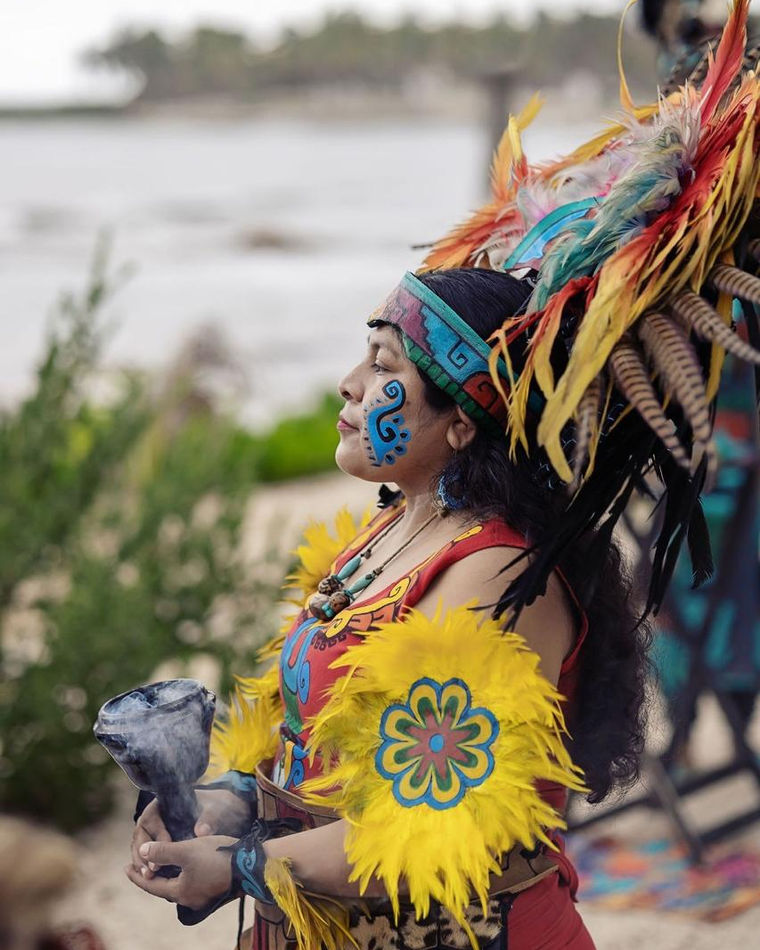 Dancer in a feathered headdress holding a smoking incense burner, with the sea behind her