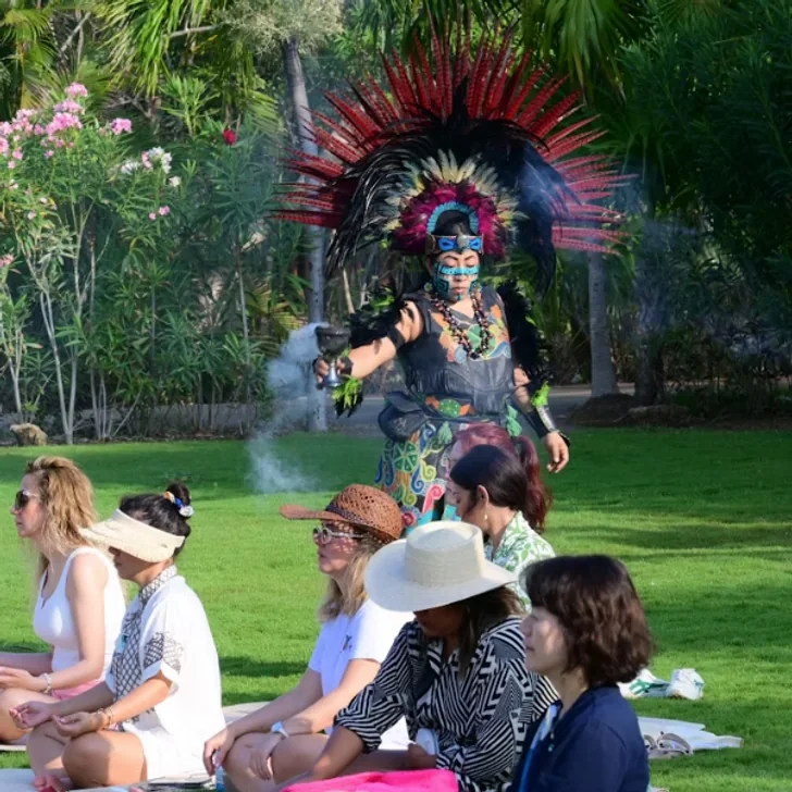 Mayan dancer with a feathered headdress before a seated group in a garden
