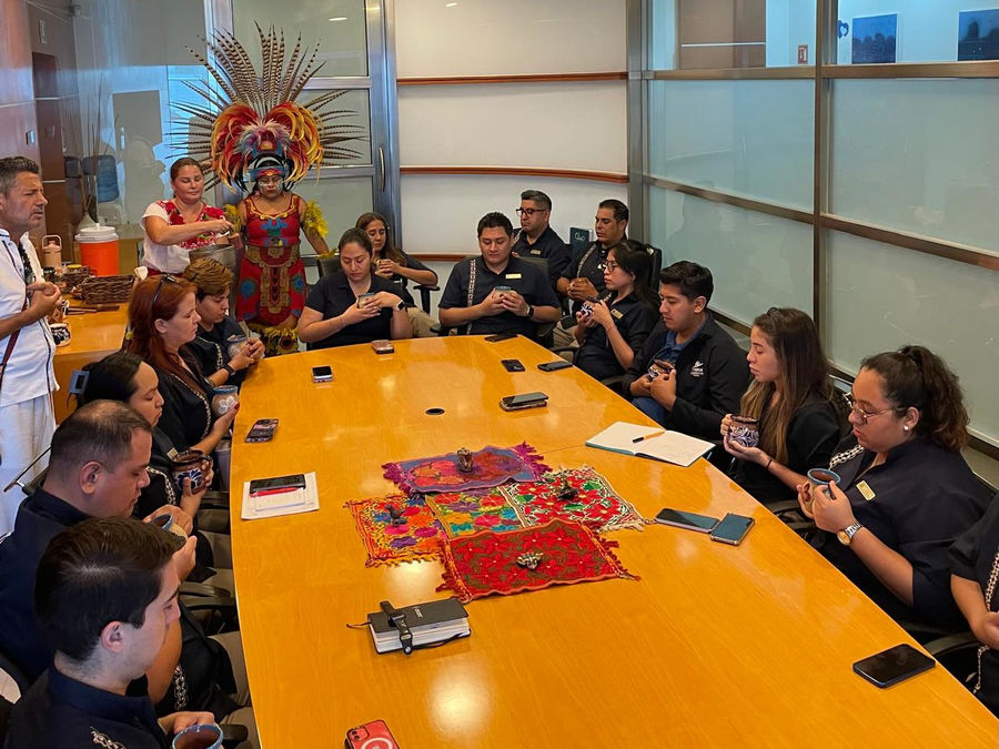 A ceremony in a meeting room: a team around a long table with an embroidered cloth, and a prehispanic dancer