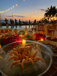  Mesa de boda al aire libre en México durante el atardecer, decorada con flores, velas y un plato de mariscos sobre hielo, con vista al mar y luces colgantes.