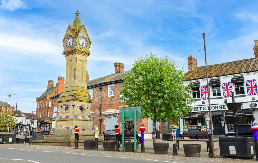 Clock tower in Thirsk town square, adorned with purple bunting. British flags hang on buildings. Clear sky, tree, and festive mood evident. Check out Crafts of Thirsk