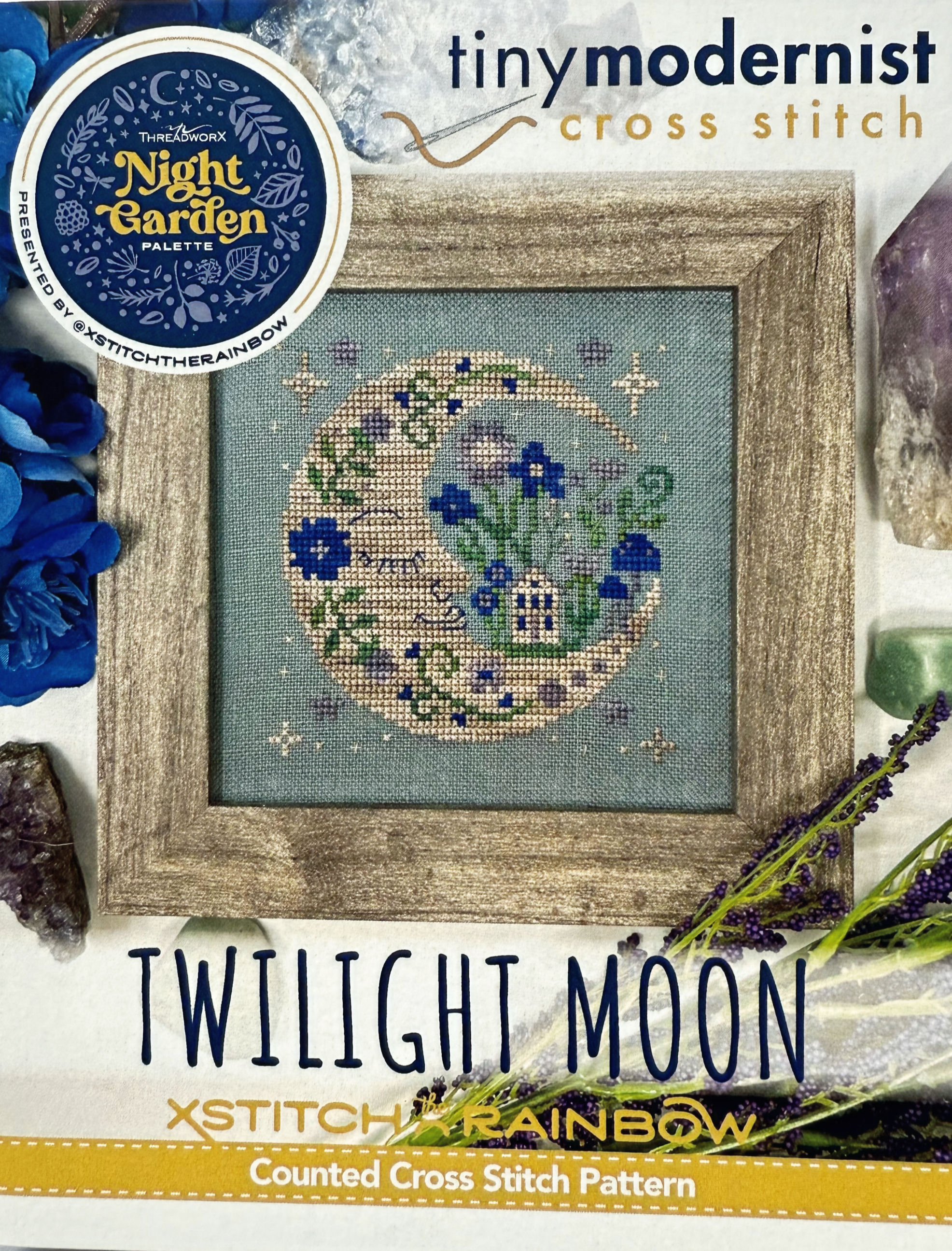 Front cover of “Twilight Moon” showing a stitched crescent moon with florals and a small house in blue tones.
