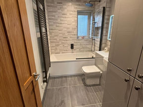 Long modern bathroom featuring grey wall tiles, bathtub, toilet and tall storage unit