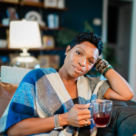 Aunty Charmaine reclining on a couch holding a glass of red tea or wine, draped in a blue plaid wrap, wearing stacked bracelets, looking directly into the camera with calm, grounded intensity in a warmly lit home setting.
