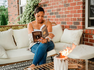 Aunty Charmaine seated on a white outdoor sofa reading a book beside a lit torch candle on a patterned patio, with a warm brick wall and ivy backdrop in a peaceful outdoor setting.