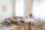 Bright living and dining area in the Erlenbad Resort with large dining table (set, flowers & candle), two upholstered chairs, beige sofa, coffee tables and floor-to-ceiling windows with transparent curtains.