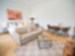 Bright living and sleeping area in the Erlenbad Resort with double bed, sofa, coffee table and carpet, plus dining area with table and four chairs; warm light from floor lamp and colored wall picture above the bed.