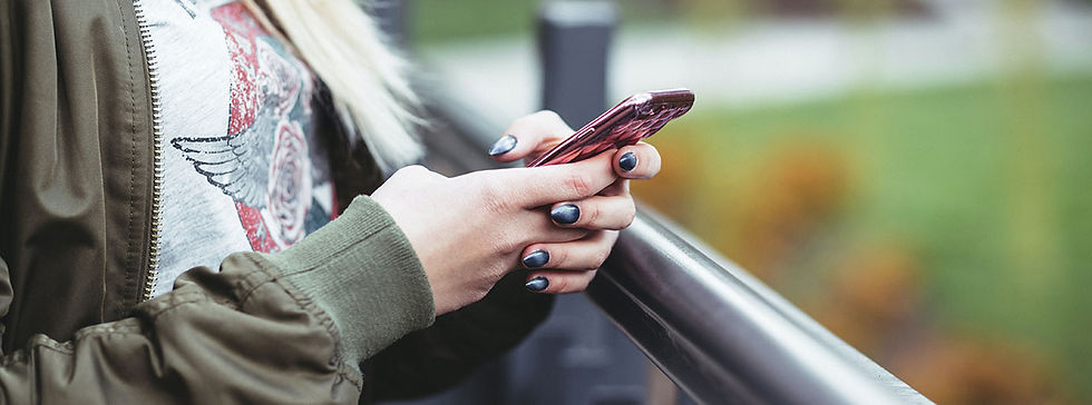Blond girl with green jacket stands by railing looking down at phone held with two hands. Image by Yura Fresh on Unsplash.