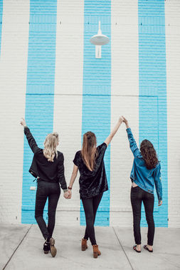 Three women hold hands and raise them in victory as they face a light blue and white striped wall. Image by Katy Ann on Unsplash.