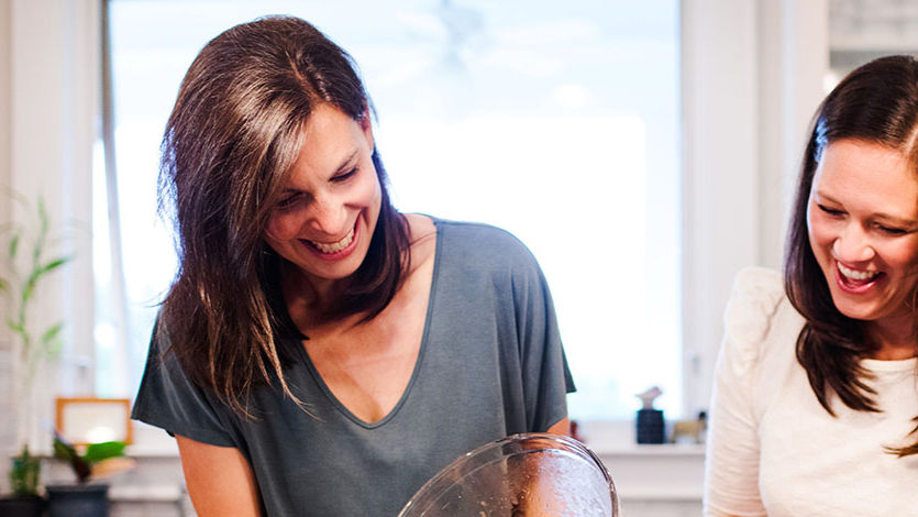 Two women with dark hair laugh together as they prepare food in the kitchen. Image courtesy of Two Alpha Gals.