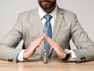Businessman in gray suit and blue tie frames a light bulb with his hands over a wooden desk, suggesting an idea.