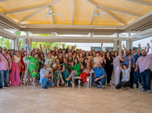 Large group of smiling people posing and waving in a bright open pavilion with yellow canopy and palm views