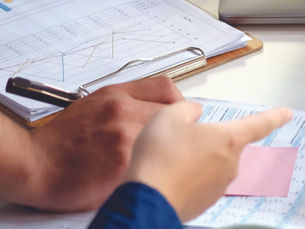 Hands point to printed charts on a clipboard at an office desk, with a coffee mug and laptop nearby.