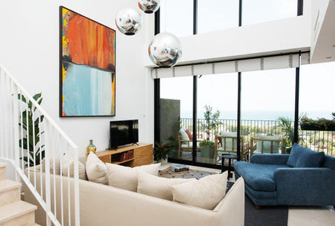 Modern living room with artwork, sofa, TV, and large windows with ocean view.