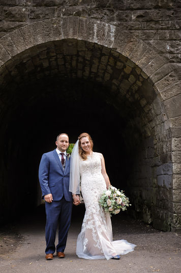 A bride in a lace gown holding a bouquet of white roses and greenery stands hand in hand with the groom in a blue suit, both smiling in front of a historic stone archway.