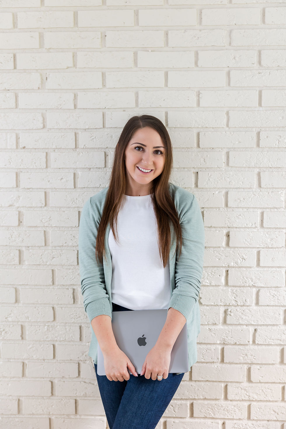 Woman in a green cardigan holding a laptop against a white brick wall
