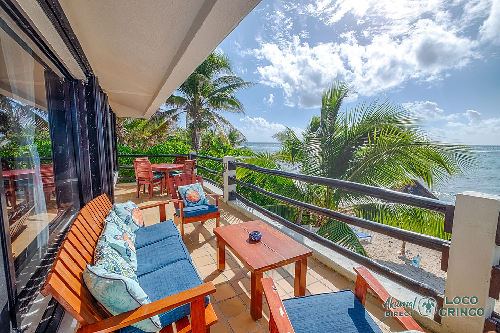 Balcony with wooden furniture and blue cushions overlooks a tropical beach. Palm trees sway under a bright, cloudy sky. Relaxing setting.