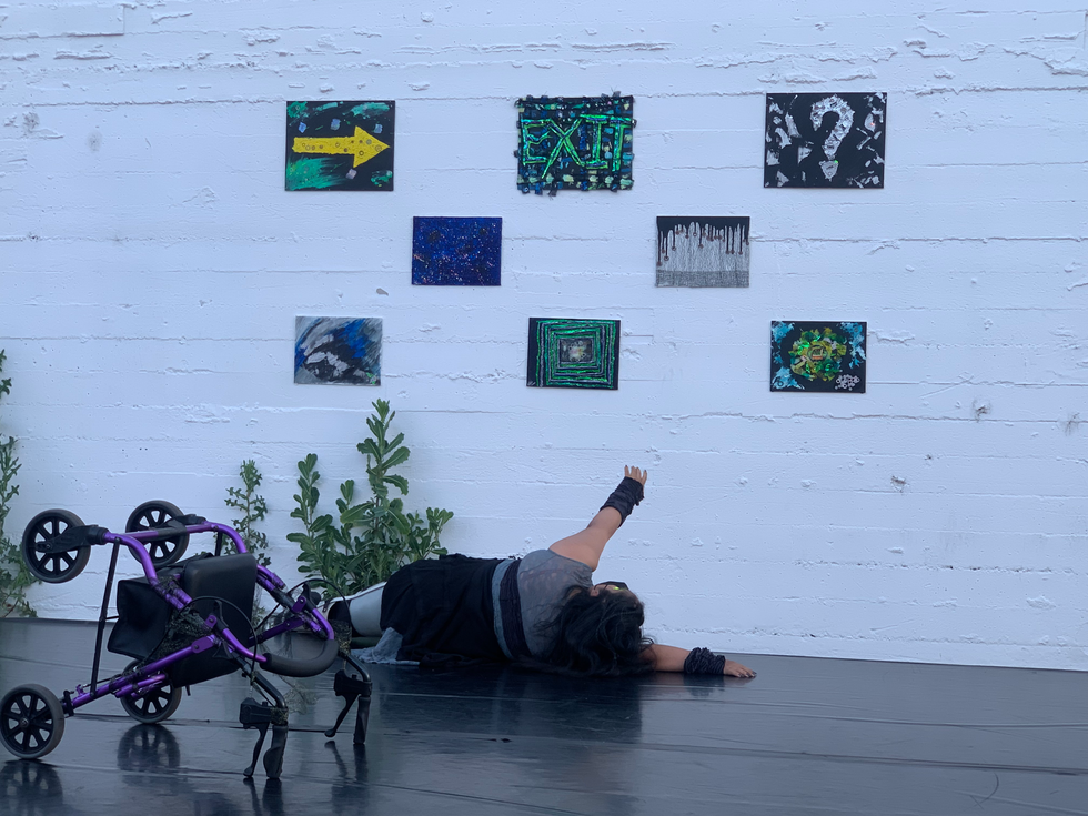 [Image description: a still image of Vanessa. She is on the floor facing all the canvases and reach up towards them. She is wearing a black & grey outfit with her hair down. The three large canvases at the top from left to right: a canvas with a yellow arrow pointing to the right. It has metal gears and green strikes behind it. The second canvas has black burlap material lined up like a graph with splats of blue & yellow ink with textured paste. In the center it says "EXIT" in 3D acrylic with neon green & chromatic colors.  The third canvas with a black background has a big 3D acrylic paint metallic silver question mark with small papers stuck that say "exit?" & has metal gears. 

The 5 smaller bottom canvases from left to right & top to bottom: the first one is a textural canvas that has blue 3D acrylic. It has glitter sprinkled all over. The second one on the top is a canvas with mesh black & gold fabric as a background. At the top it has black acrylic paint dripping down with metal gears at the bottom of each paint drip. The bottom first canvas has streaks of blue, black, & silver acrylic paint. The second canvas is a rectangular maze constructed out of blue burlap lace giving it a 3D effect. In the center it has a small rectangle filled with 3D transparent acrylic paint. Inside the acrylic is a paper that says "Exit?" with small gears. The last canvas has  a small wooden TV with an empty cut out as a screen. Surrounding it are scraps of burlap painted with green, yellow, & white ink. In the corners are streaks of textured paste with light blue ink.]