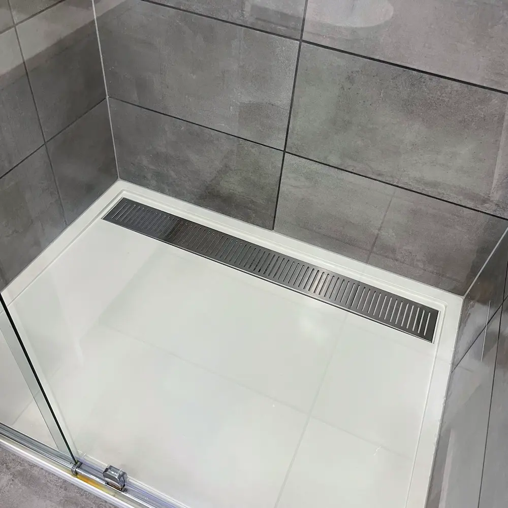 Walk-in shower enclosure featuring descaled grey wall tiles and polished stainless steel linear trench drain by Whole Detail. 
