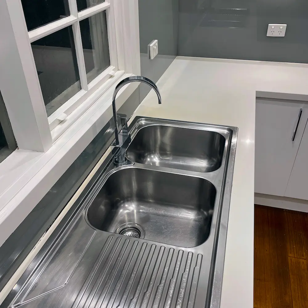 Dual-bowl stainless steel kitchen sink, polished mixer tap, and white window frame detailed by Whole Detail. 