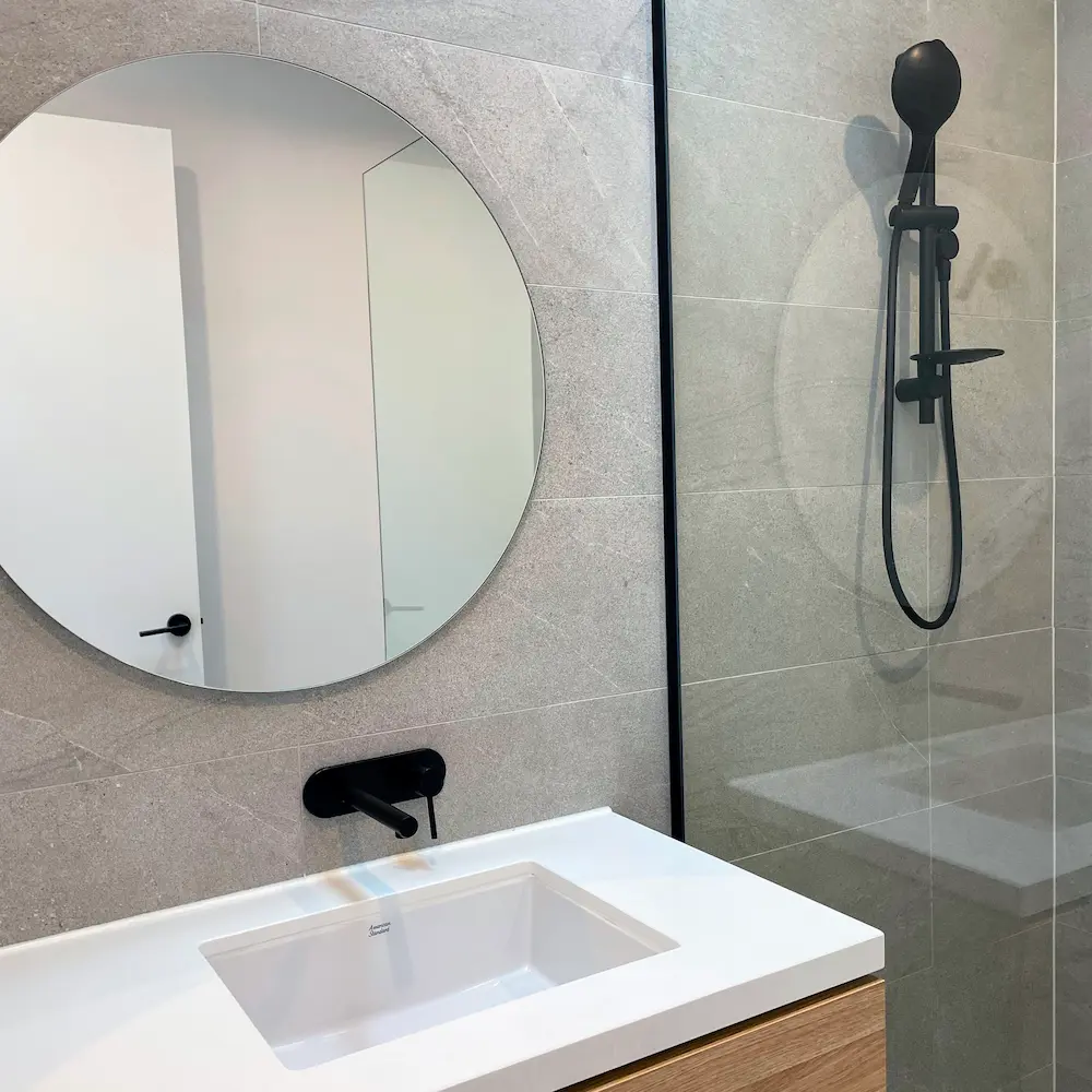 Ensuite bathroom suite with polished circular mirror, stone vanity top, and descaled matte black shower fixtures by Whole Detail. 