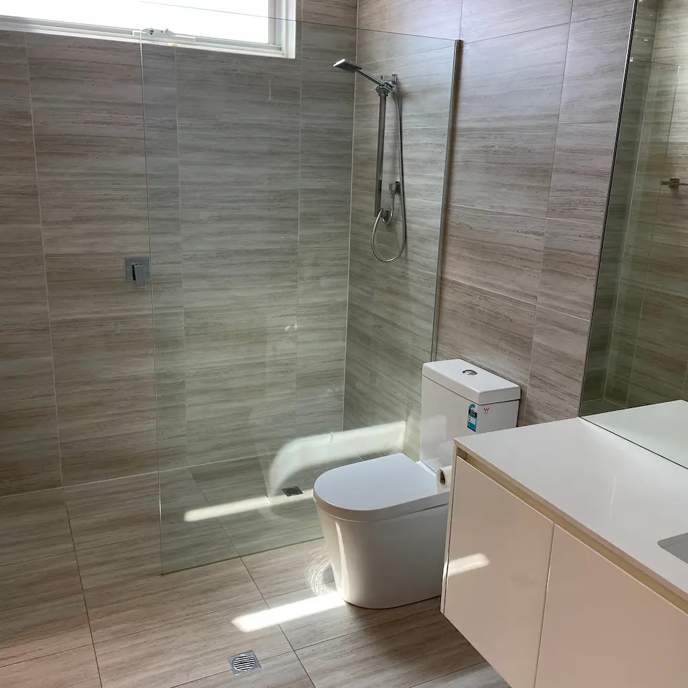 Ensuite bathroom featuring descaled frameless glass shower panel, sanitized toilet, and neutral stone-look wall tiles. 