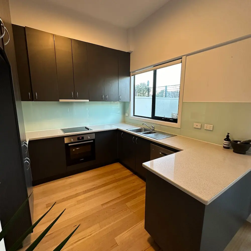  Modern L-shaped kitchen with dark cabinetry, glass splashback, and polished timber floors detailed by Whole Detail. 