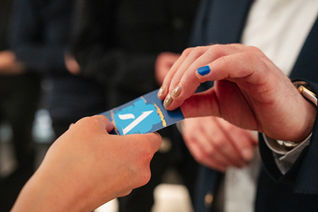 Nahaufnahme einer geschäftlichen Visitenkartenübergabe bei einem Networking-Event. Eine Person im dunkelblauen Anzug mit blauer Nagellackfarbe auf einem Finger nimmt oder gibt eine blaue Visitenkarte mit weißem Symbol weiter. Die Szene deutet auf ein professionelles Business-Meeting oder eine Veranstaltung hin, mit unscharfem Hintergrund und weiteren Teilnehmern in dunkler Kleidung.